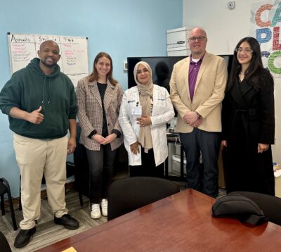 Five people pose for a photo in an office setting. A whiteboard with a list of names is on the wall behind them, and a wooden conference table is in the foreground.