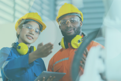 Two engineers in safety gear and helmets stand together indoors. One holds a tablet and gestures while the other listens, both wearing protective eyewear and yellow ear protection.