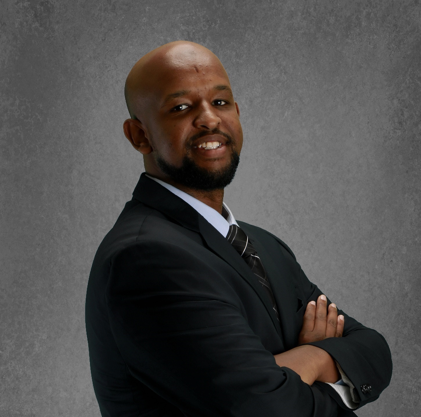 A man in a black suit and striped tie stands with arms crossed, smiling, in front of a gray textured background.