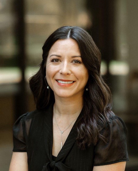 Woman with long dark hair and a black blouse smiling at the camera, standing in front of a blurred indoor background.