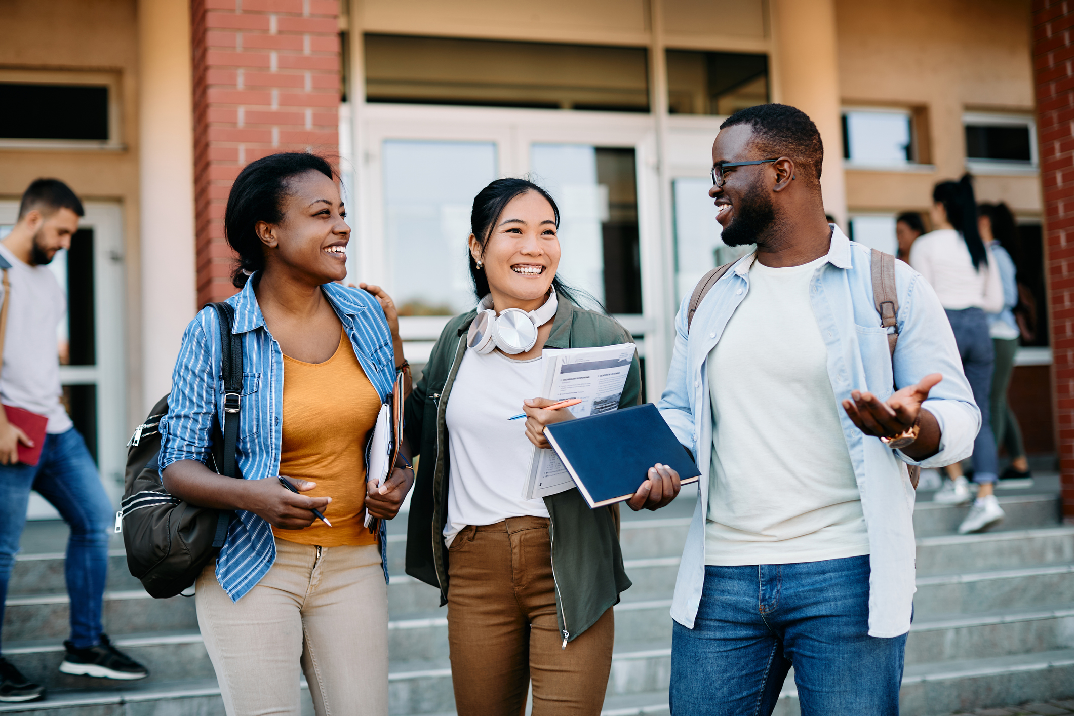 Three students walk together outside a building, smiling and talking while holding notebooks and papers. Other people are visible in the background on the steps.