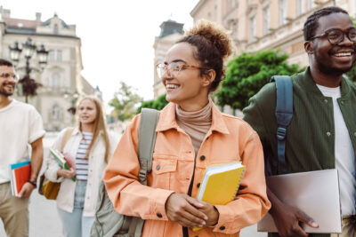 Four young adults walk outdoors in a city, smiling and carrying books and a laptop, with historic buildings and trees in the background.