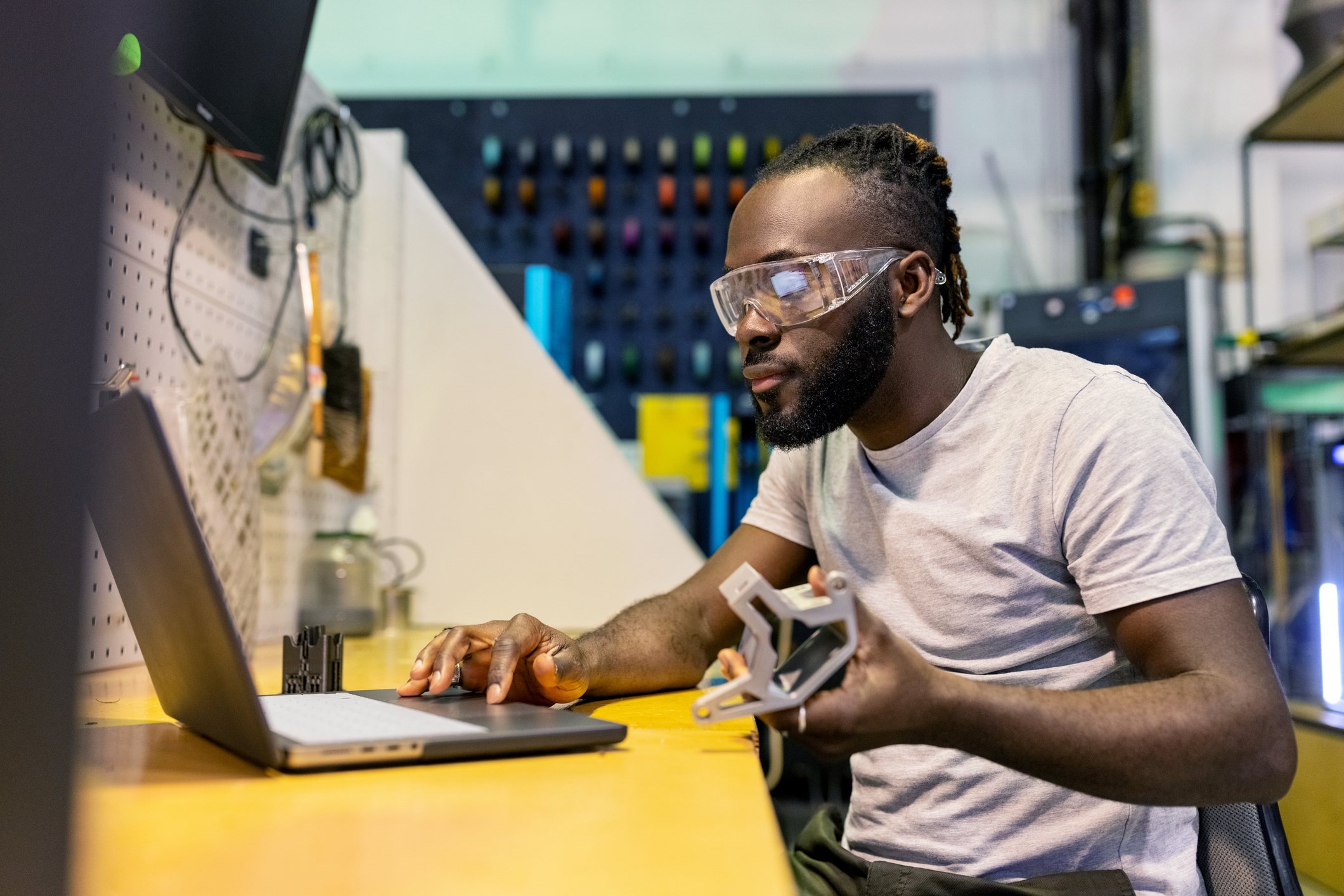 A man wearing safety glasses works on a laptop at a desk, holding a 3D-printed object in his left hand in a workshop setting.
