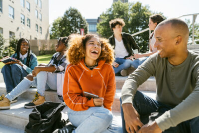 A group of young adults sits on outdoor steps, talking and laughing together on a sunny day, with buildings and trees in the background.