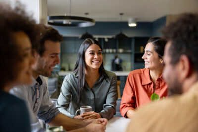 Five people sit around a table, smiling and talking in a bright, modern kitchen or office space. One woman in the center holds a coffee mug.