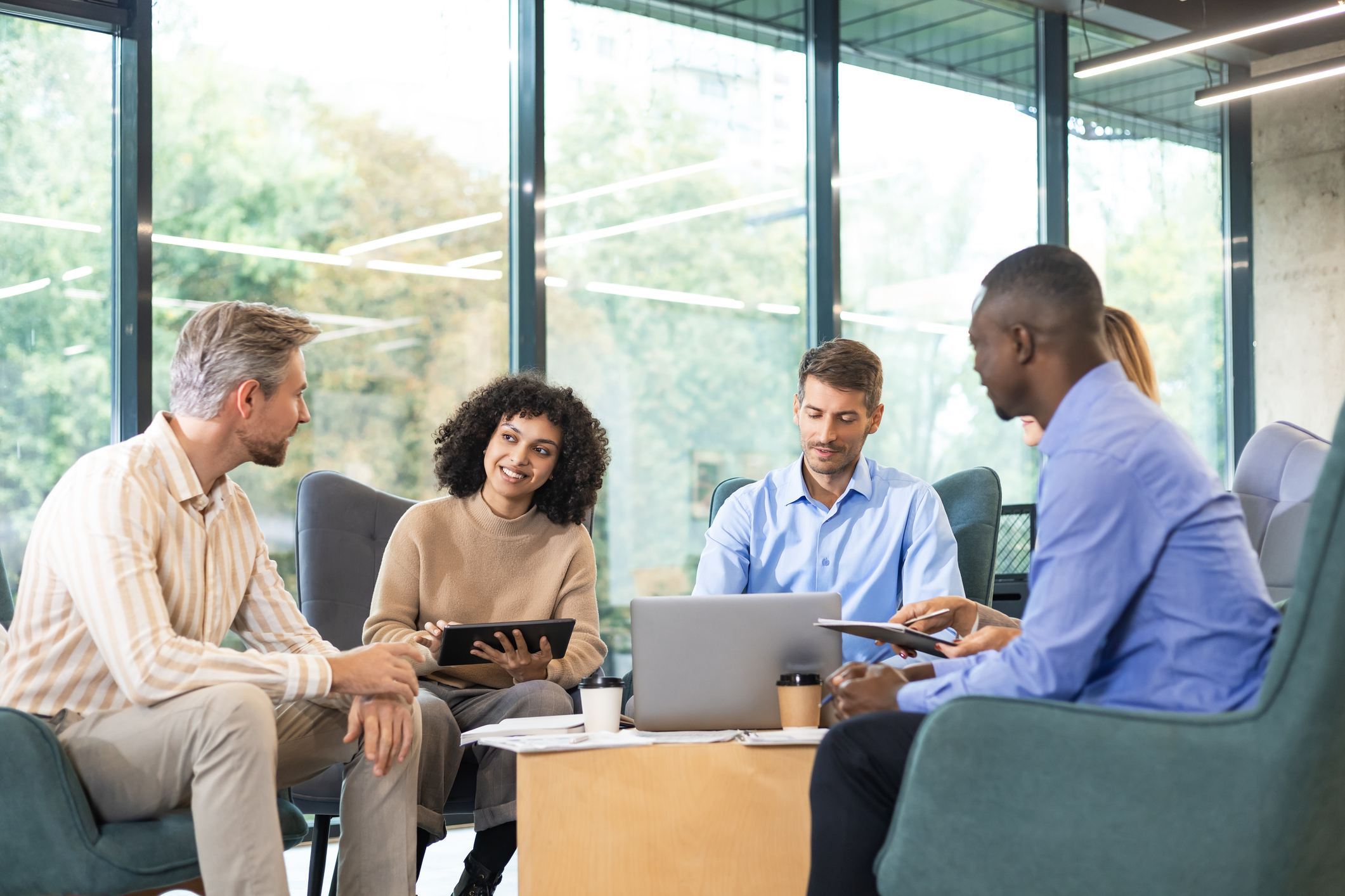 Five people sit in modern office chairs around a wooden table, using laptops and tablets, engaged in a discussion in a bright, glass-walled meeting room.