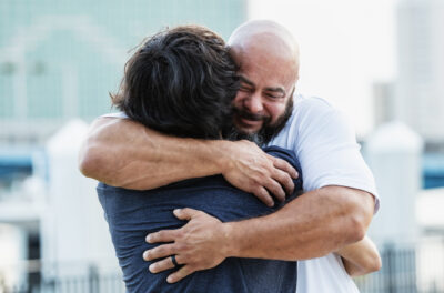 Two men embrace tightly outdoors, one appearing emotional with closed eyes and a slight smile. Tall buildings and a blurred city background are visible.