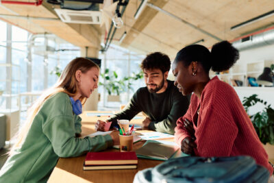 Three people sit at a table in a bright, modern office, discussing documents and using colored pens. Books, a cup, and a pencil holder are on the table.