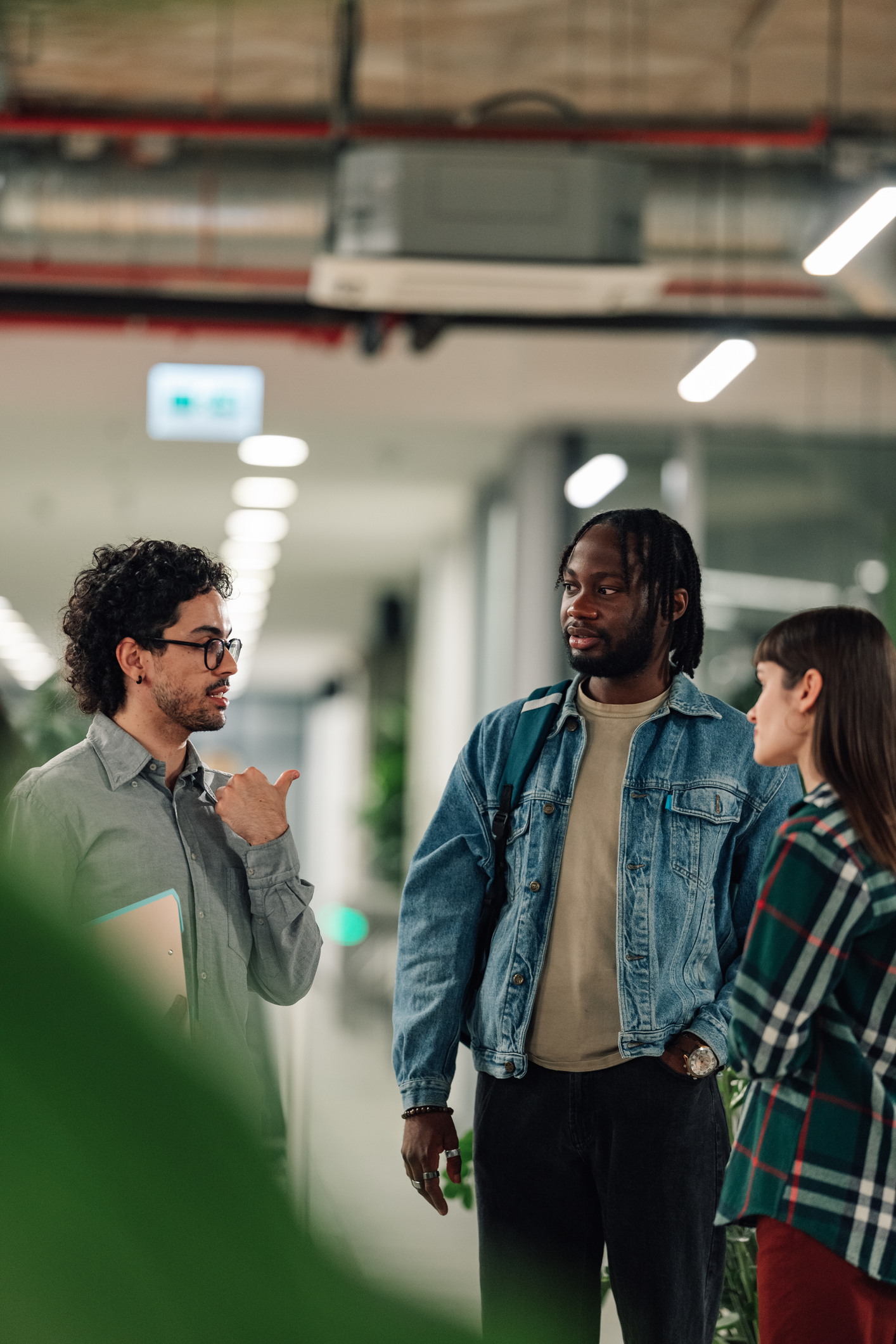 Three people stand in a hallway having a conversation, with one person gesturing with his hand while the others listen.
