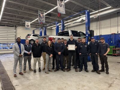 A group of auto shop employees stands together in a garage, with one person in the center holding a framed certificate. A pickup truck is lifted on a car lift behind them.