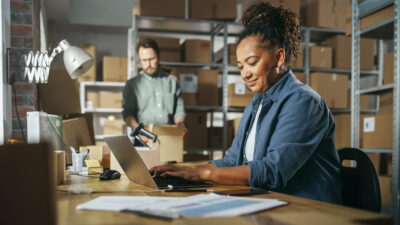 A woman works on a laptop at a desk in a warehouse while a man scans a box in the background. Shelves filled with boxes are visible throughout the space.