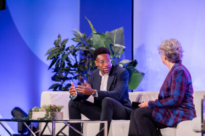 Two people sit on a white couch engaged in conversation on a stage, with green plants and a blue-lit background behind them.