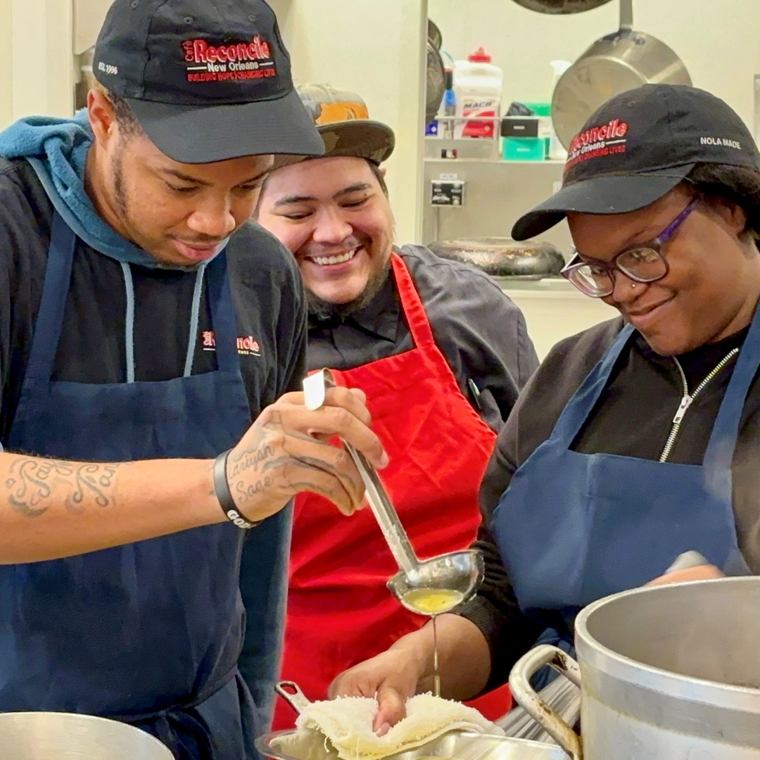 Three people in aprons and hats work together in a kitchen, with one person ladling soup into a bowl while the others observe and smile.