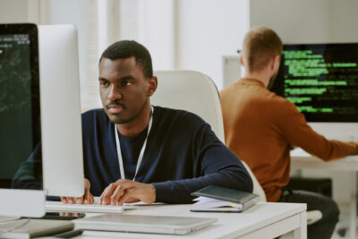 Two men work at computers in a modern office; one faces the camera, the other faces away with code displayed on his monitor.