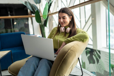 Woman in a green sweater and headphones sits in a chair using a laptop, with plants and modern decor in the background.