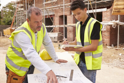 Two people in safety vests review building plans at a construction site; one points at the plans while the other takes notes on a clipboard. Scaffolding and bricks are visible in the background.
