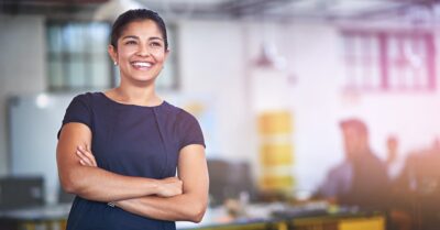 Woman in a navy dress stands with arms crossed and smiles in a modern office setting, blurred coworkers and desks in the background.