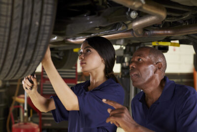 Two mechanics in blue uniforms work under a raised vehicle, with one person using a tool and the other pointing and giving instructions.