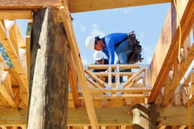 Two construction workers wearing safety gear install wooden beams on a building frame under a clear sky.
