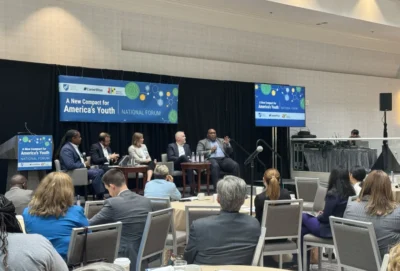 A panel of five speakers sits on stage at a conference titled “A New Compact for America’s Youth,” with an audience seated and listening attentively.