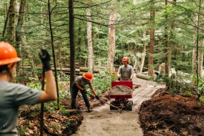 Three workers in orange helmets build a trail in a forest, with one shoveling gravel, one pushing a wheelbarrow, and one standing in the foreground holding a tool.
