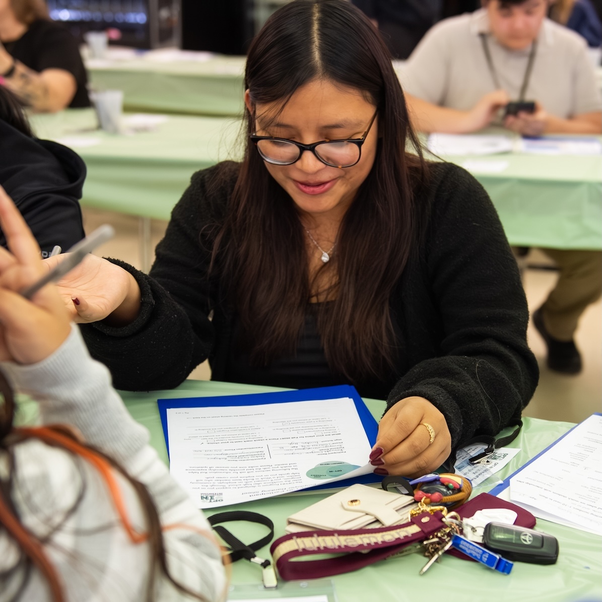 A woman with glasses reviews documents at a table, holding a pen. Keys, a phone, and a wallet are on the table beside her. Other people are present in the background.