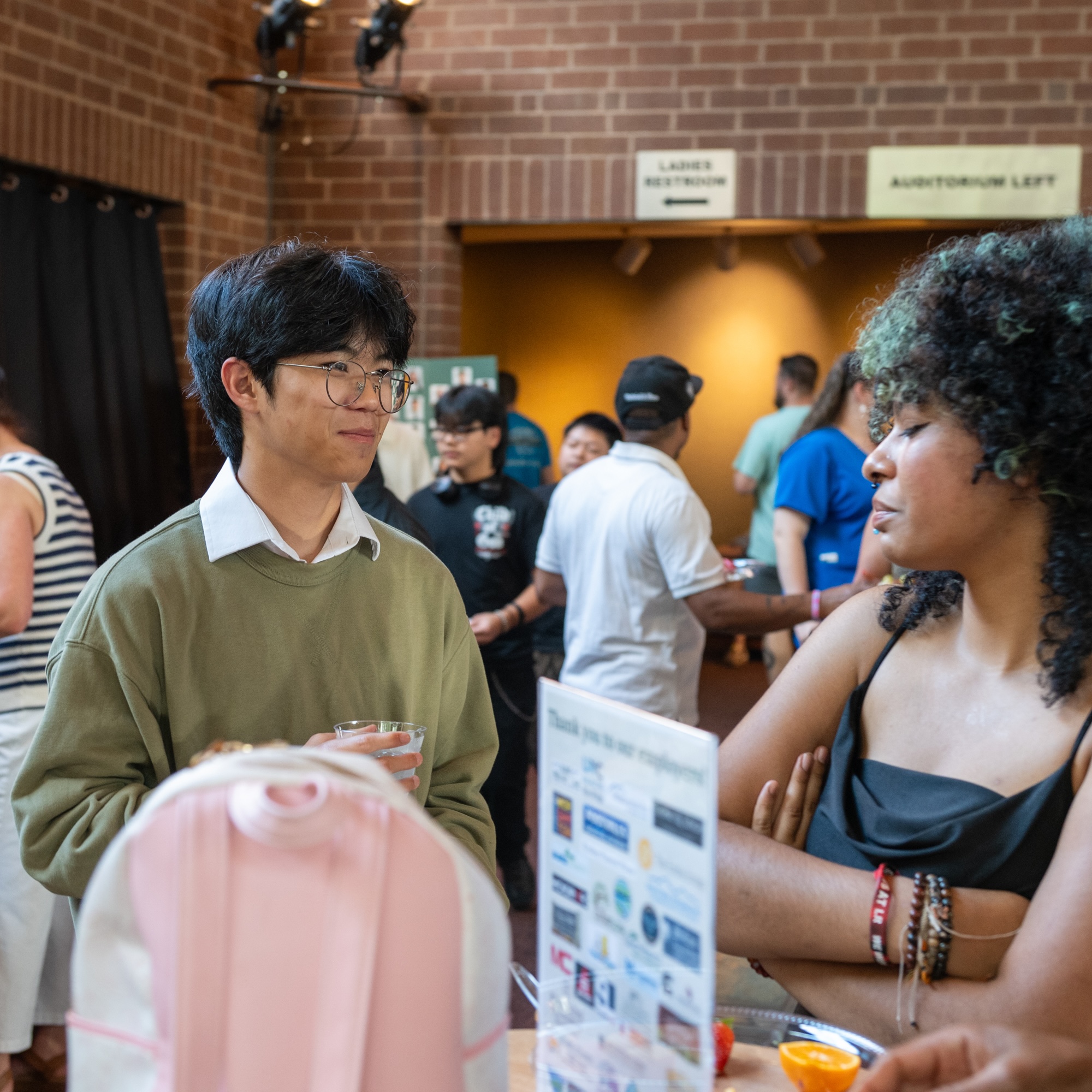 Two people stand and talk at an indoor event near a table with a sign and drinks. Other attendees are visible in the background.