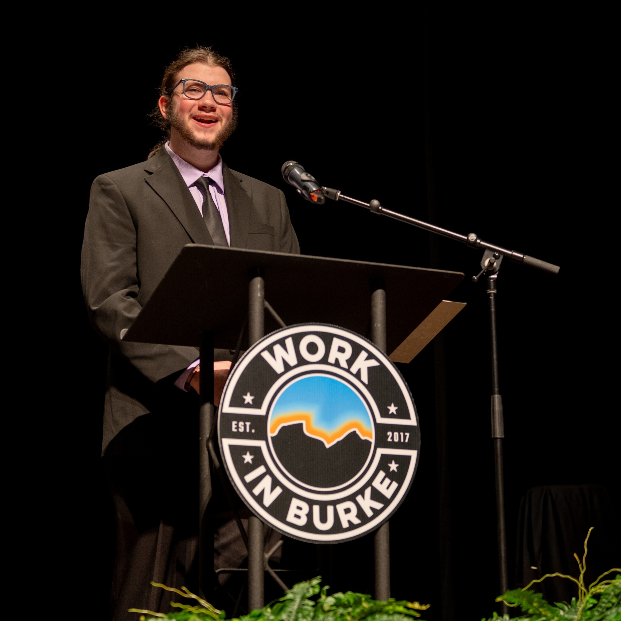 A person in a suit stands at a podium with a “Work in Burke” sign, speaking into a microphone on a stage.