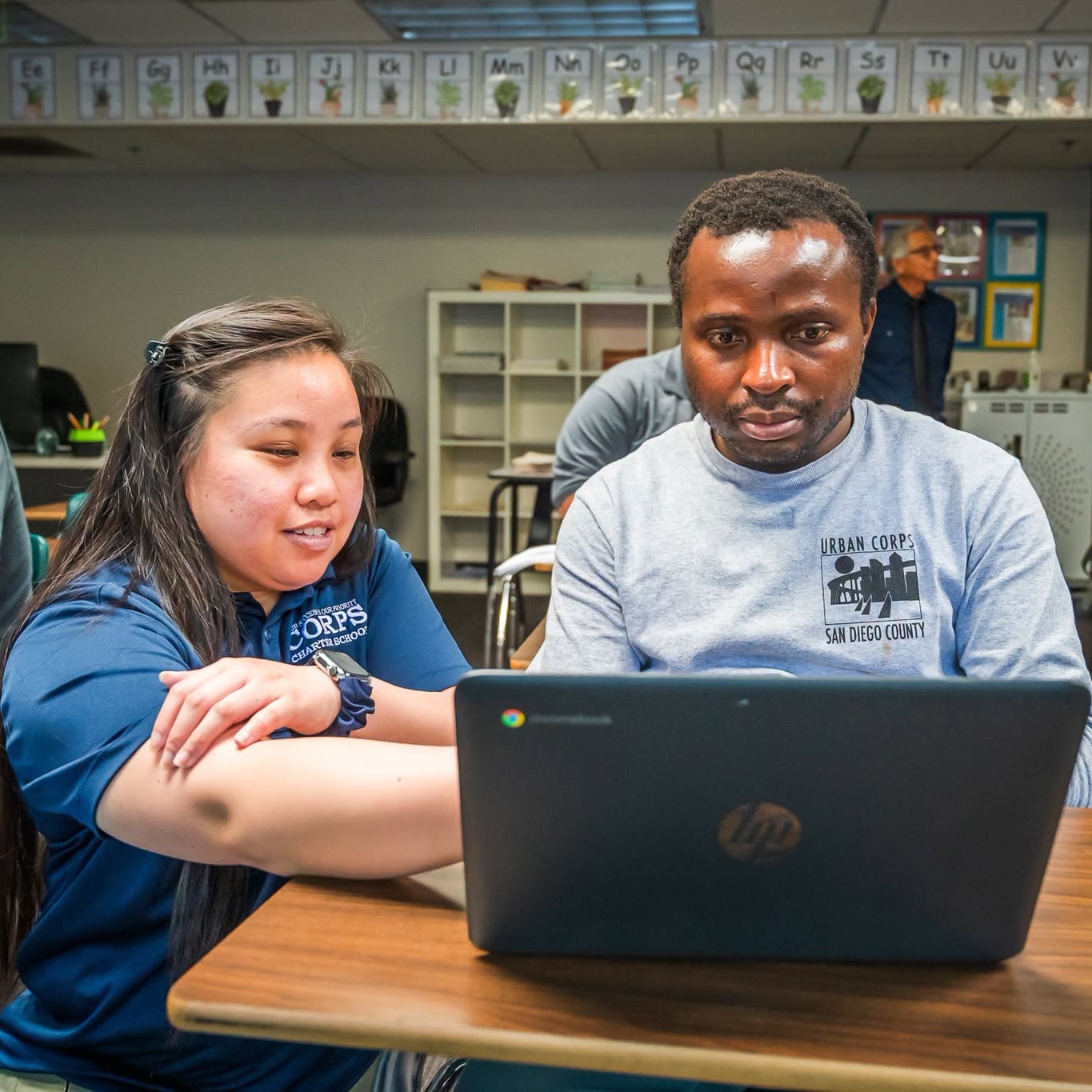 Two people sit at a table, looking at a laptop screen together in a classroom setting, with alphabet cards displayed along the wall behind them.