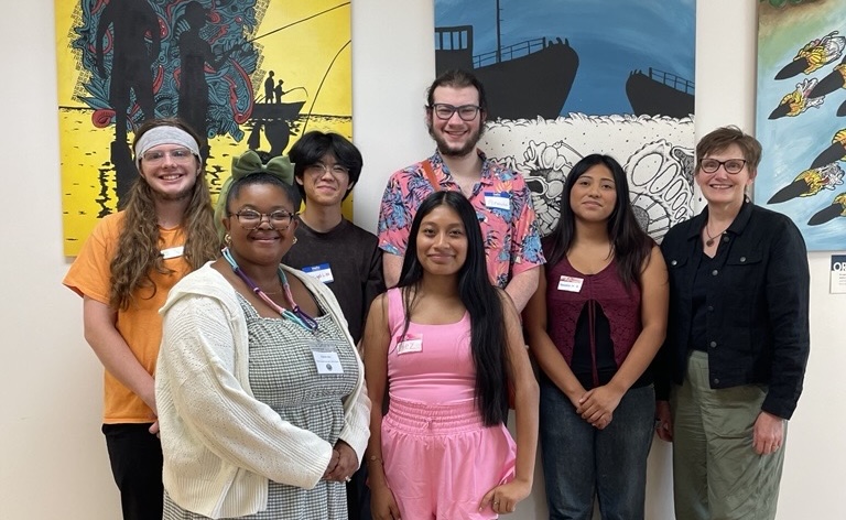 Seven people stand in front of colorful artwork, posing for a group photo. Most wear name tags and are smiling.