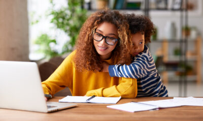 Woman in a yellow sweater working on a laptop with notebooks nearby while a young child hugs her from behind.