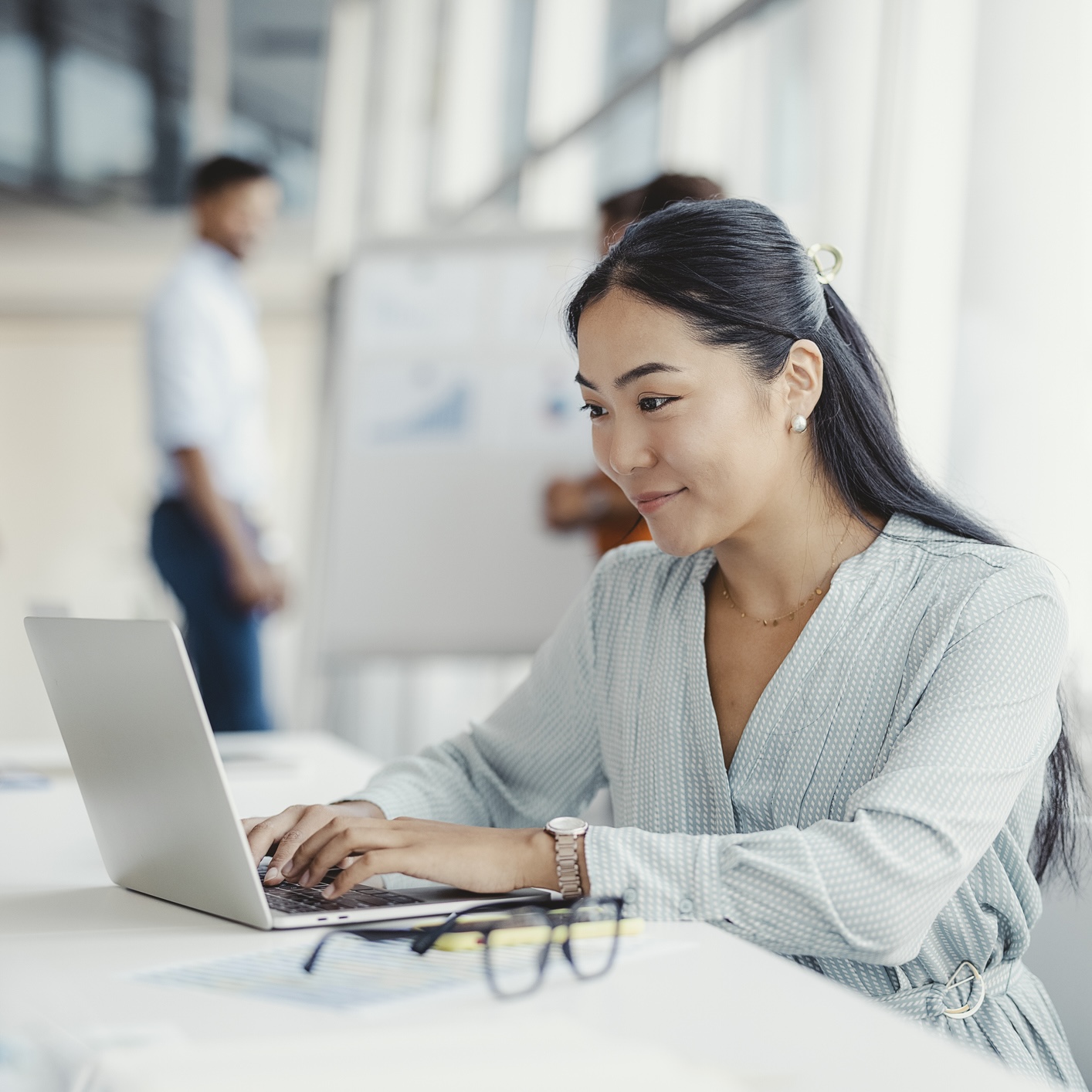 Woman typing on a laptop at a desk in a bright office, with two people conversing in the background near a whiteboard.