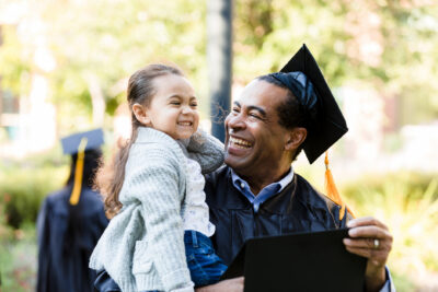 A man in a graduation cap and gown holds a smiling young girl outdoors, both appearing happy.