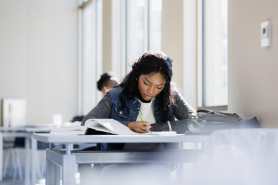 A woman sits alone at a table by a window, writing in a notebook with an open textbook in front of her.