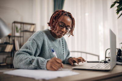Person wearing glasses and a gray sweater writes on paper while sitting at a desk with a laptop in a bright, home office setting.