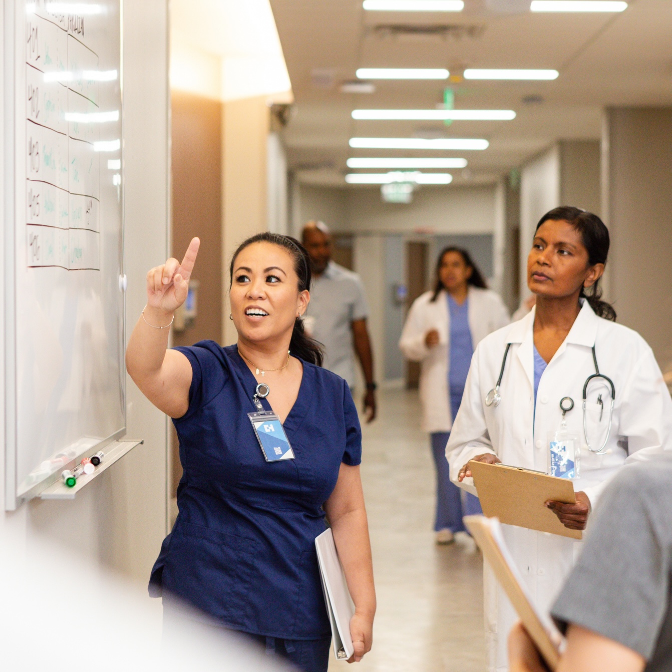 A nurse points at a whiteboard while a doctor holding a clipboard watches in a hospital hallway. Other medical staff are in the background.