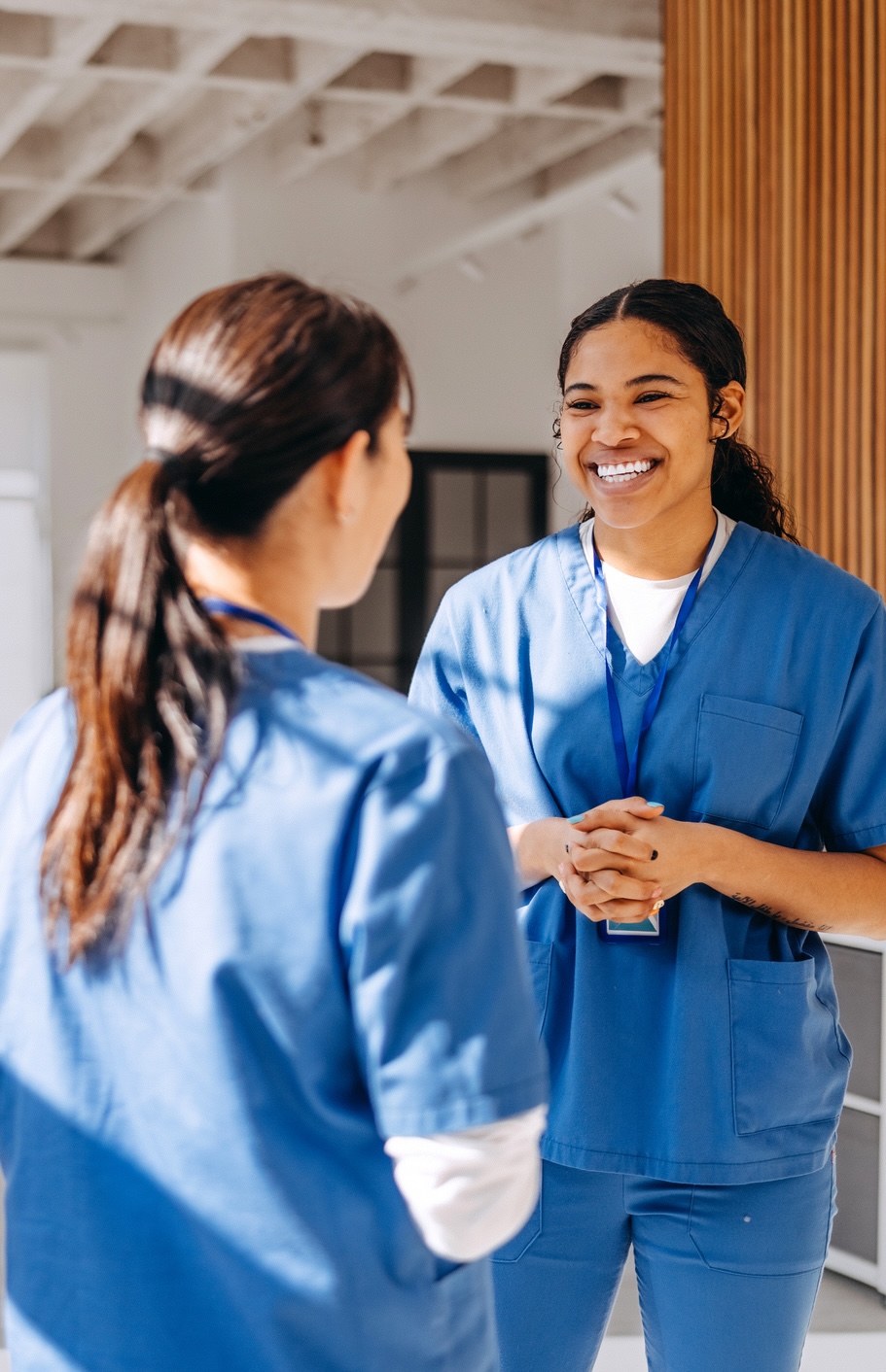 Two healthcare workers in blue scrubs stand indoors, facing each other and talking. One is smiling and holding her hands together. Both wear lanyards and have their hair pulled back.