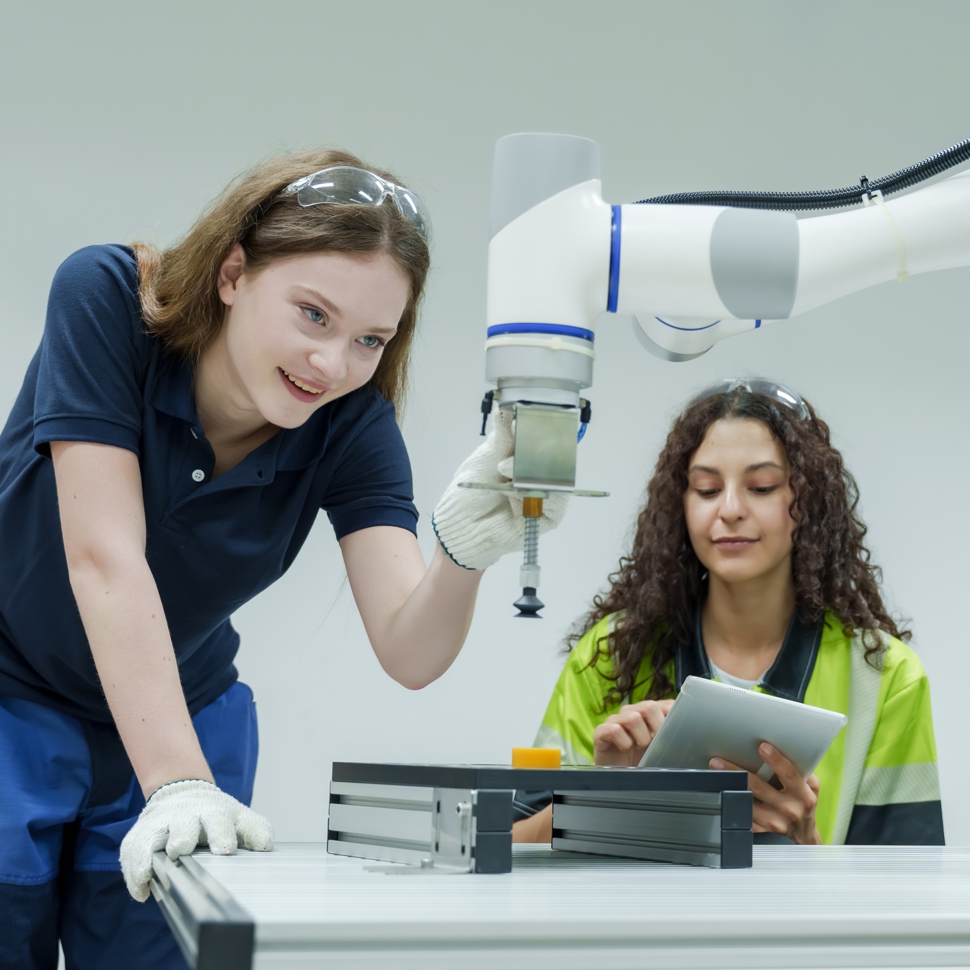 Two women work with a robotic arm at a table. One adjusts the arm, while the other holds a tablet, observing and possibly recording data. Both wear safety gear.