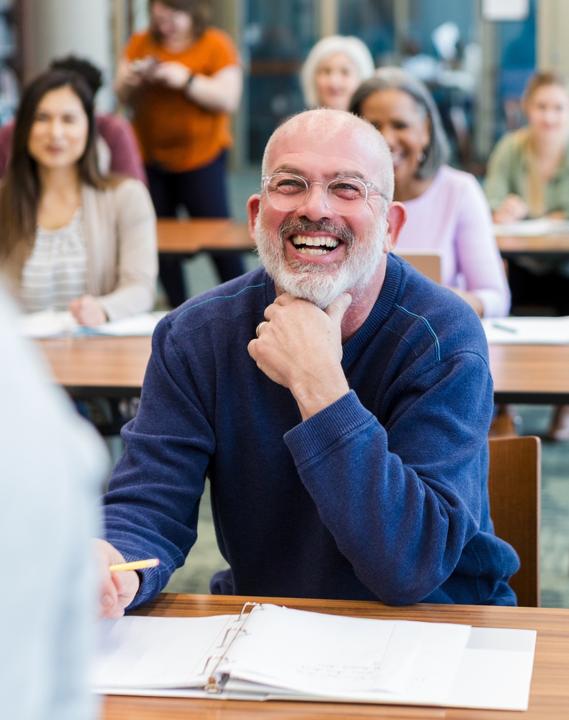 A smiling man with glasses and a beard sits at a desk in a classroom, holding a pencil and facing the instructor. Other adults are seated and attentive in the background.