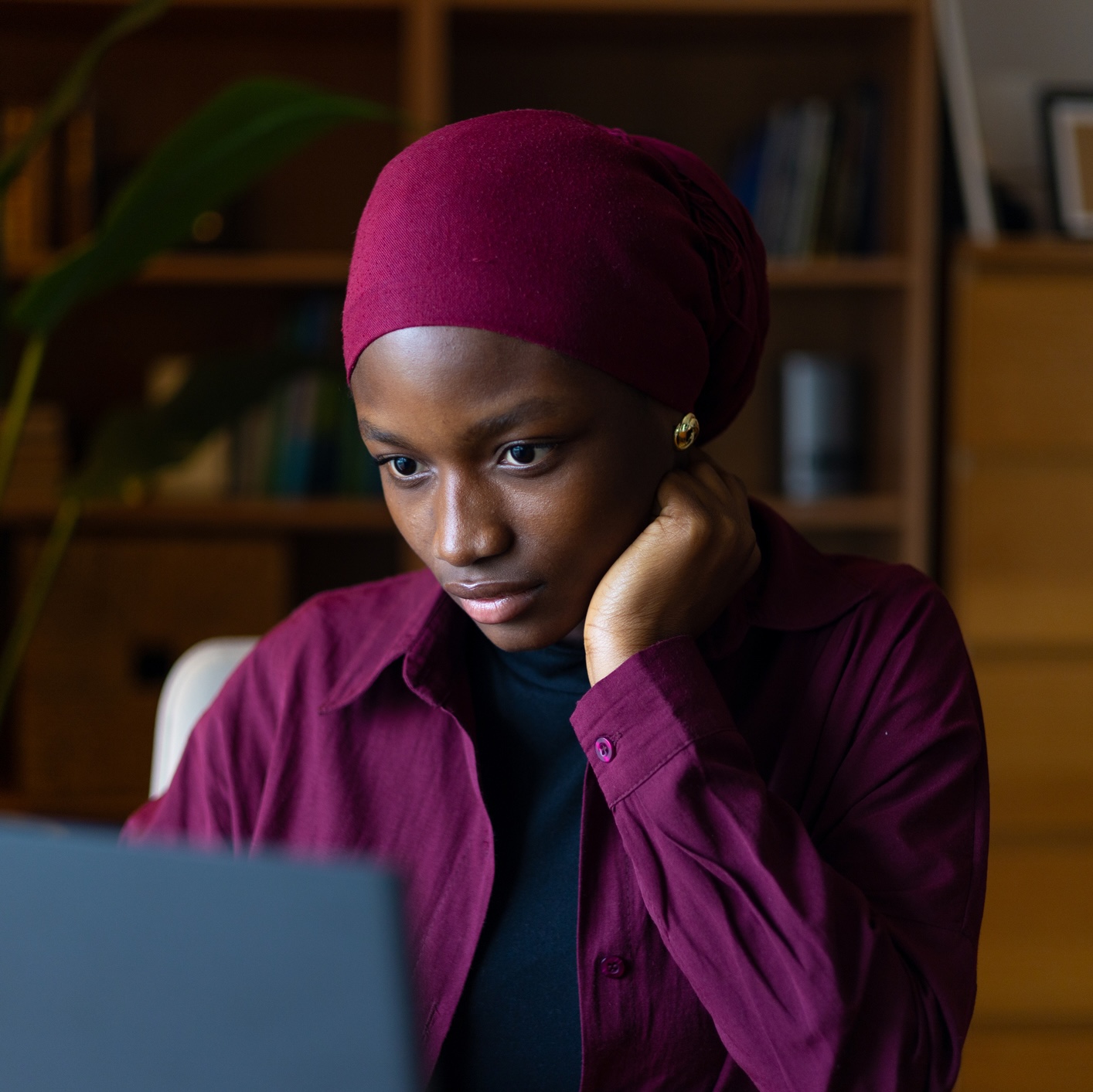 A woman in a burgundy headscarf sits indoors, looking intently at a laptop screen with her hand resting on her cheek.