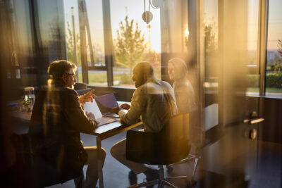 Three people sit around a conference table in a glass-walled office, talking and using a laptop, with sunlight streaming in through the windows.