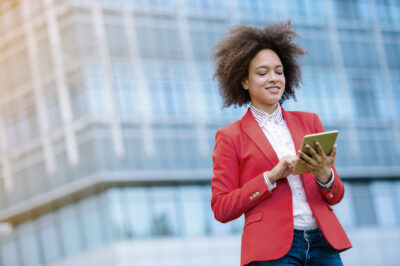 A person in a red blazer stands outside a glass building, looking at and using a tablet.