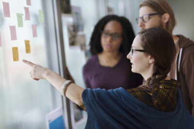 Three people stand by a window covered with sticky notes. One person points to a note while the others observe, suggesting a group discussion or planning session.