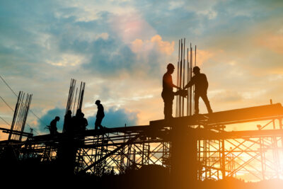 Silhouetted construction workers stand and work on scaffolding at sunset, with metal rods extending upwards against a cloudy sky.