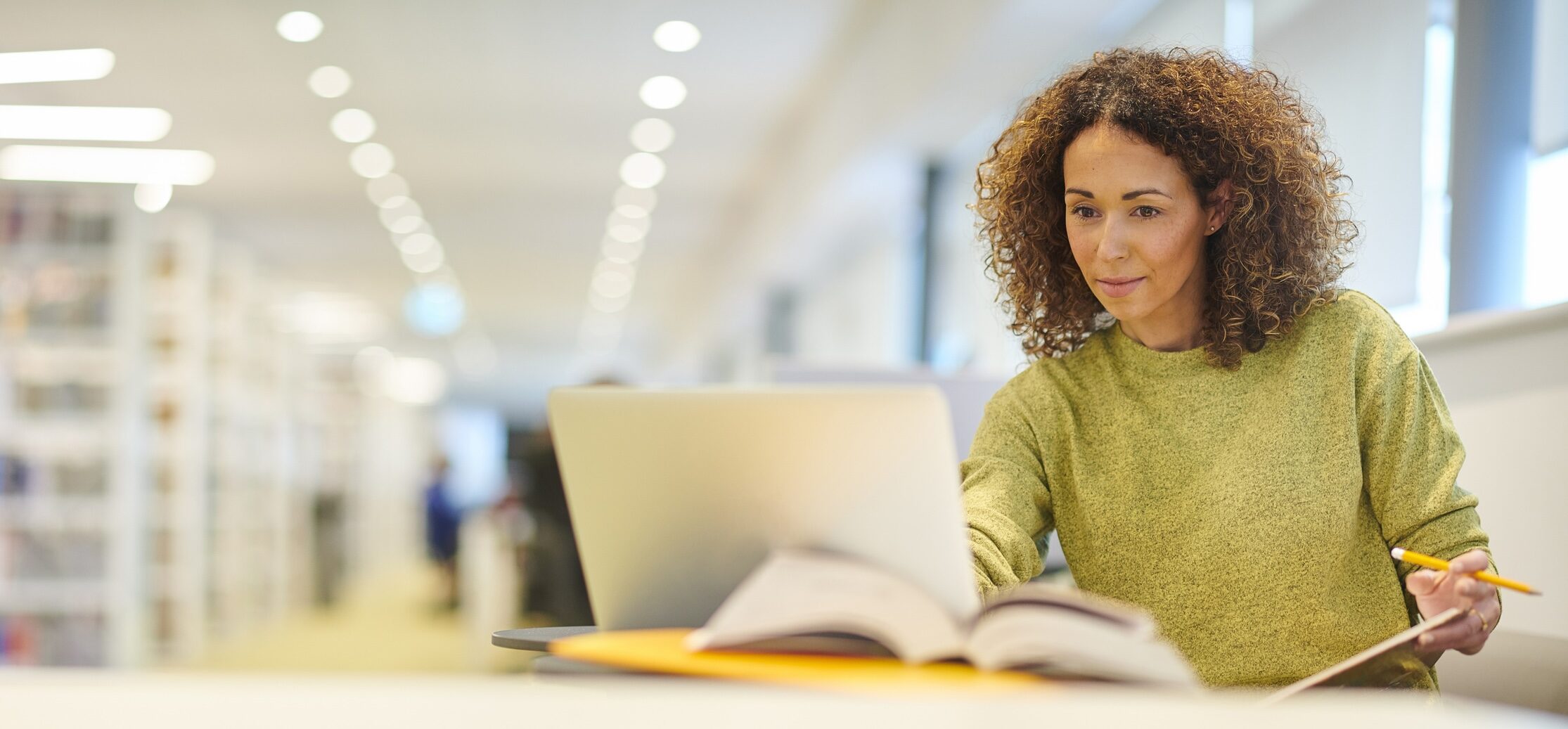 A woman with curly hair studying at a laptop and open book in a library.