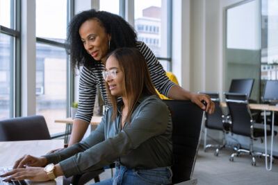 Two women collaborating at a desk in an office, with one woman typing at a computer while the other leans over her shoulder to look at the screen.