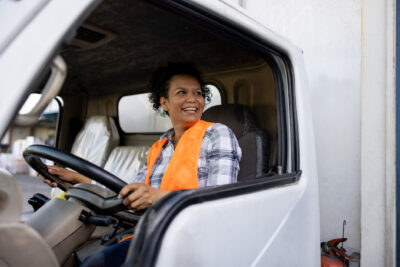 A smiling woman in an orange safety vest sitting in the driver’s seat of a truck.