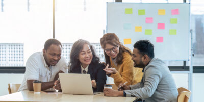 Four colleagues collaborate around a laptop at a table in a bright office, with a whiteboard featuring sticky notes in the background.