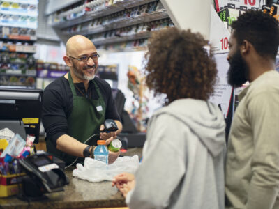 A smiling convenience store clerk scans a beverage for two customers at the checkout counter.