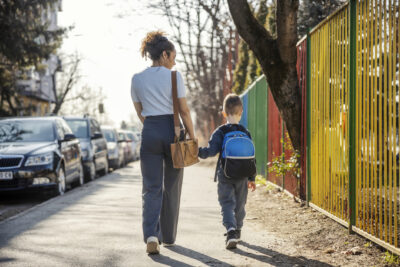 A woman and a young boy with a backpack holding hands while walking down a sidewalk beside a colorful fence.